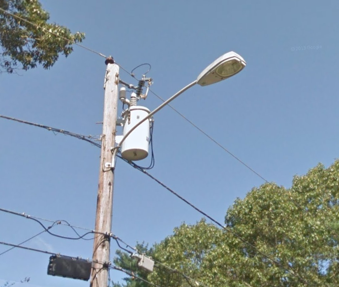100W MV GE M-250A2 FCO
In North Kingstown, RI. This one is a 1984-1987 model. NECo used a LOT of these in the 80s and sadly removed a lot of them less than 10 years later when the HPS onslaught occurred. The utility pole is pre-1965 too since it's got a brown insulator instead of gray. 

The transformer has been replaced though. They went around and replaced most of the transformers around 15-20 years ago. They replaced more old ones around 5 or so years ago after a transformer blew in Johnston, RI and fried appliances in several homes on the small dead-end street. Of course NGrid didn't pay the homeowners anything to replace their TVs, computers, microwaves, refrigerators, toasters, coffee makers, and whatever else was fried by the transformer blowing. I'm glad I wasn't one of those residents. Another reason to dislike LEDs since they're all circuit board powered and would fry too.
Keywords: American_Streetlights