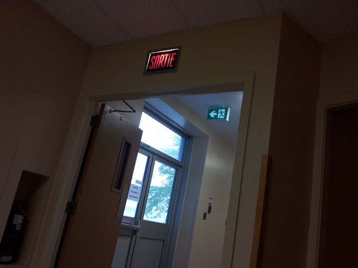 Transition has begun!
The new running-man exit sign was installed last summer during a partial remodeling in my school. For some reason the old one hasn't been removed, so now this exit is identified by both the existent and the new standard!

The older SORTIE seems to have fluorescents now, probably a PL retrofit kit. The brand new unit is very likely LED.
Keywords: Indoor_Fixtures